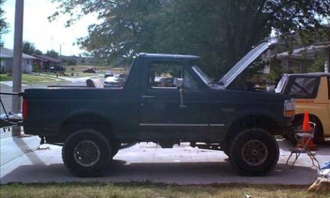 1995 Ford Bronco - Driver-side profile of a dark green 1990s Ford Bronco with the rear hardtop removed and the hood raised, parked in a residential driveway.…
