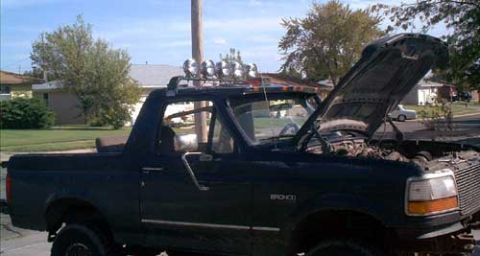 1995 Ford Bronco - Dark green 1992-96 Ford Bronco parked at a curb with the hood raised and the hardtop removed, shot from the driver's side. A roll bar carri…