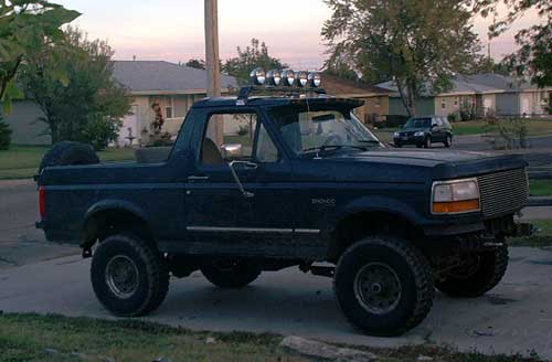 Dark blue lifted Ford Bronco, top off, with roof light bar at a curb