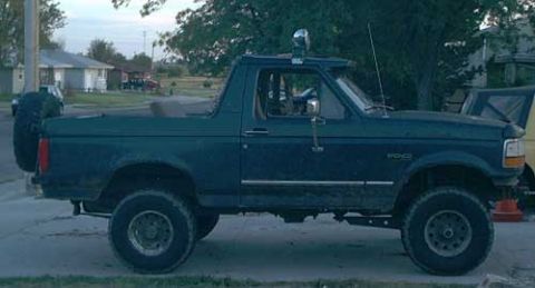 1995 Ford Bronco - Driver-side profile of a dark green 1990s Ford Bronco with the rear top removed, showing the roll bar with a mounted spotlight and a CB ant…