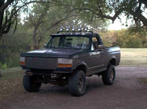 1995 Ford Bronco - A dark-colored 1990s Ford Bronco full-size SUV parked on a dirt path, shown from the front three-quarter angle with a chrome grille, lifted…