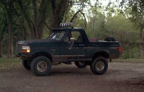 1995 Ford Bronco - Driver-side view of a dark green lifted Ford full-size Bronco with the hardtop removed, fitted with a roll bar carrying a row of off-road d…
