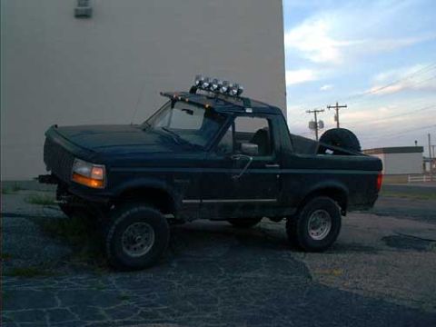 1995 Ford Bronco - Dark green lifted full-size SUV with the hardtop removed, shot from the driver's side in a paved lot at dusk. A tubular roll bar carries a…
