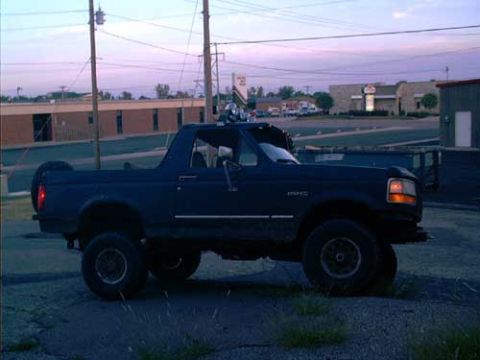 1995 Ford Bronco - Side profile of a dark blue lifted full-size Ford Bronco with its hardtop removed and a roll bar carrying off-road lights, parked in a pave…
