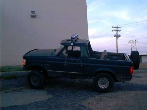 1995 Ford Bronco - Side view of a dark blue lifted Ford Bronco with the fiberglass rear top removed, showing a roll bar with a row of four round off-road ligh…
