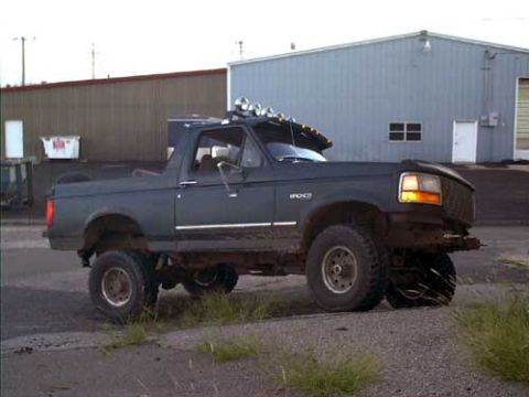 1995 Ford Bronco - Passenger-side view of a dark blue Ford Bronco with the hardtop removed, parked on gravel beside a metal shop building. It runs a lift with…