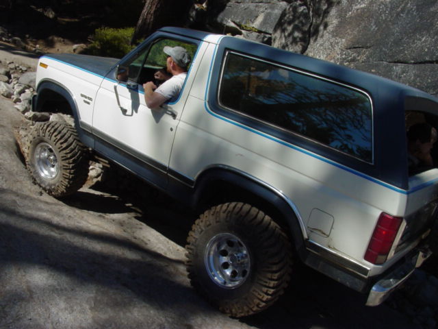 White and blue Ford Bronco with large tires climbing a rocky hill. Driver wearing a cap is visible