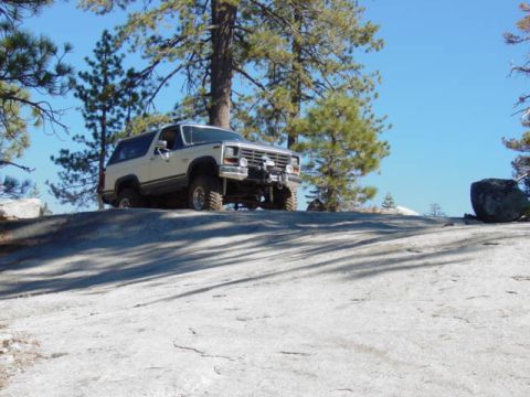 1986 Ford Bronco - This SUV is navigating a steep, rocky incline under a bright blue sky, surrounded by pine trees.