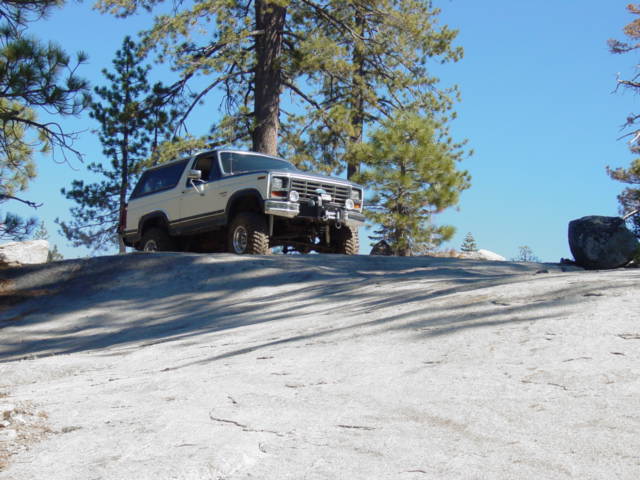 White SUV climbing a large rock formation with pine trees and a clear blue sky in the background