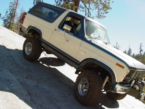 1986 Ford Bronco - Ford Bronco navigates a steep, rocky trail in a wooded area under a clear blue sky.