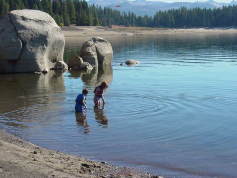 1986 Ford Bronco - Children explore the shallow waters of a lake, with large boulders and a forested shoreline under a clear sky.