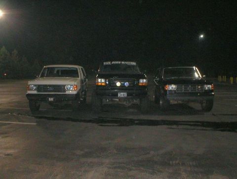 1989 Ford Bronco - Three Ford Broncos are parked in a lot at night. The middle vehicle has a banner on its windshield.