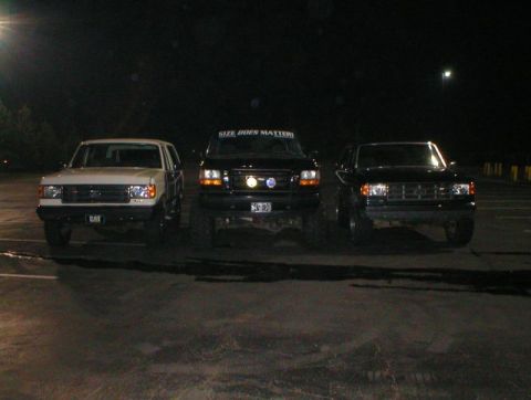 1989 Ford Bronco - Three Ford trucks are parked in a lot at night, with one displaying a humorous message on its visor.