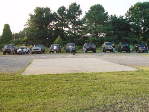 1989 Ford Bronco - Multiple SUVs and pickup trucks are parked in a line on a gravel lot with trees in the background.