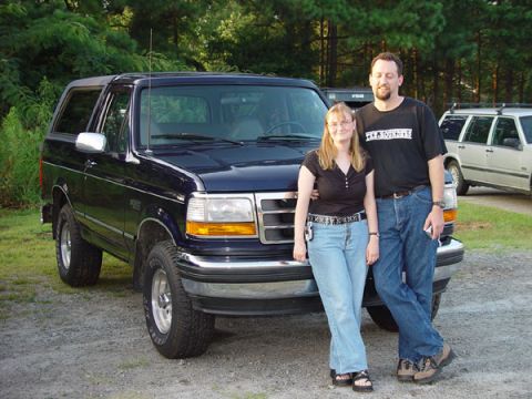 1989 Ford Bronco - A couple poses with a dark blue SUV, with another vehicle visible in the background.