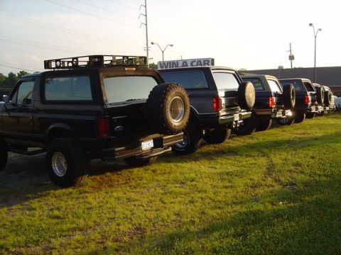 1989 Ford Bronco - A line of Ford Broncos, including a black 1989 model with a roof rack and spare tire, parked on a grassy lot.