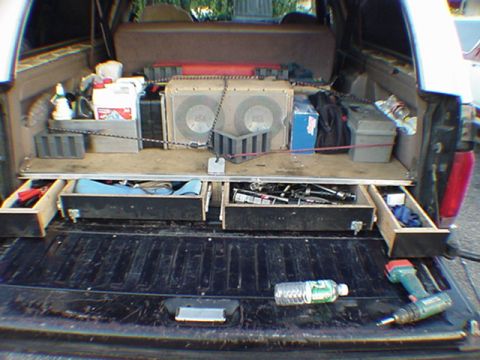 1993 Ford Bronco - Rear cargo area of an SUV with the tailgate down, showing a custom wood drawer/storage system holding two large subwoofer speakers, tools,…