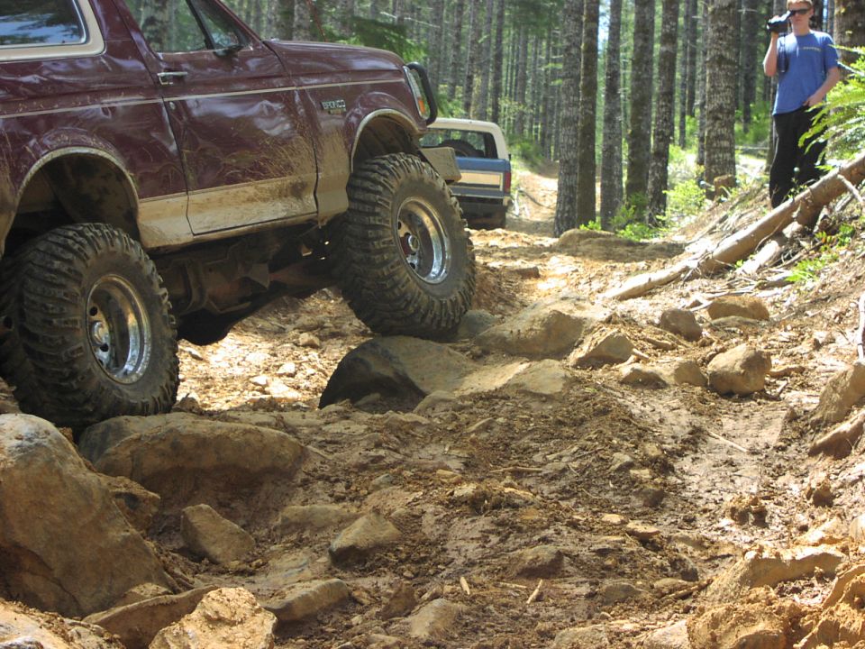 Maroon 1993 Ford Bronco climbing a rocky trail, another Bronco behind it, person filming in background