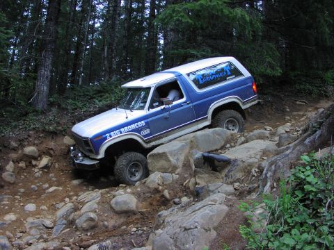 1993 Ford Bronco - A blue and white Ford Bronco tackles a challenging rocky path in a wooded area.