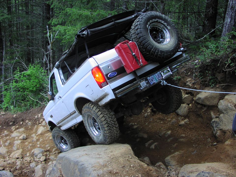 1993 Ford Bronco with soft top, spare tire, and gas can, climbing over rocks on a forest trail