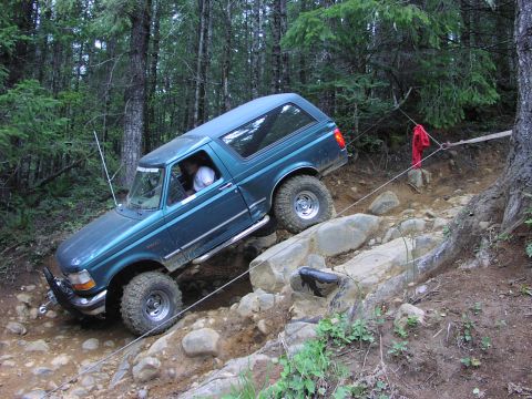1993 Ford Bronco - A teal 1993 Ford Bronco navigates a challenging rocky obstacle on an off-road trail.