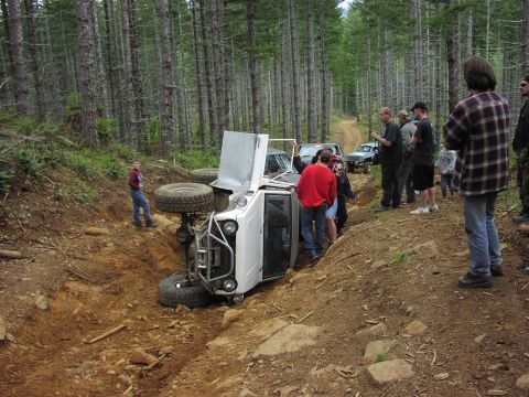 1993 Ford Bronco - A group of people observe a white off-road SUV that has rolled onto its side on a wooded trail.