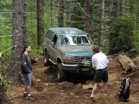 1993 Ford Bronco - A green Ford Bronco navigates a challenging off-road trail with two individuals assisting.