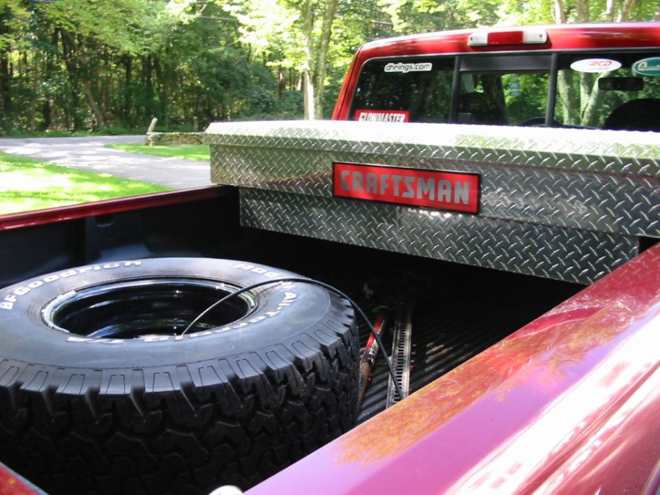 Red pickup truck bed interior showing a spare tire and a Craftsman toolbox. Trees and a road are visible in the background