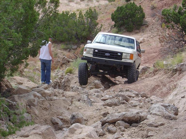 White Ford pickup descending a steep rocky wash as a person stands to the side