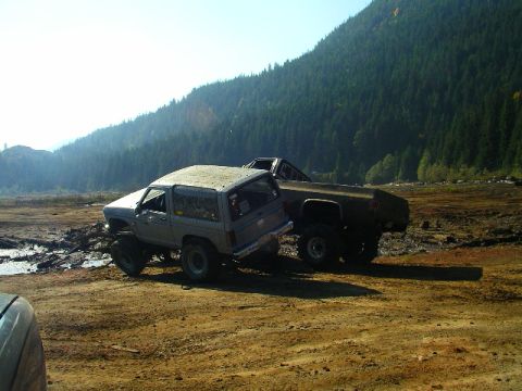 1984 Ford Bronco II - Two modified SUVs navigate a muddy terrain with a forested mountain backdrop.