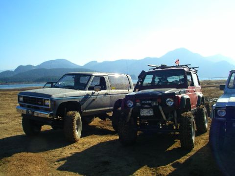 1984 Ford Bronco II - Two off-road SUVs are parked on a beach with a body of water and mountains behind them.