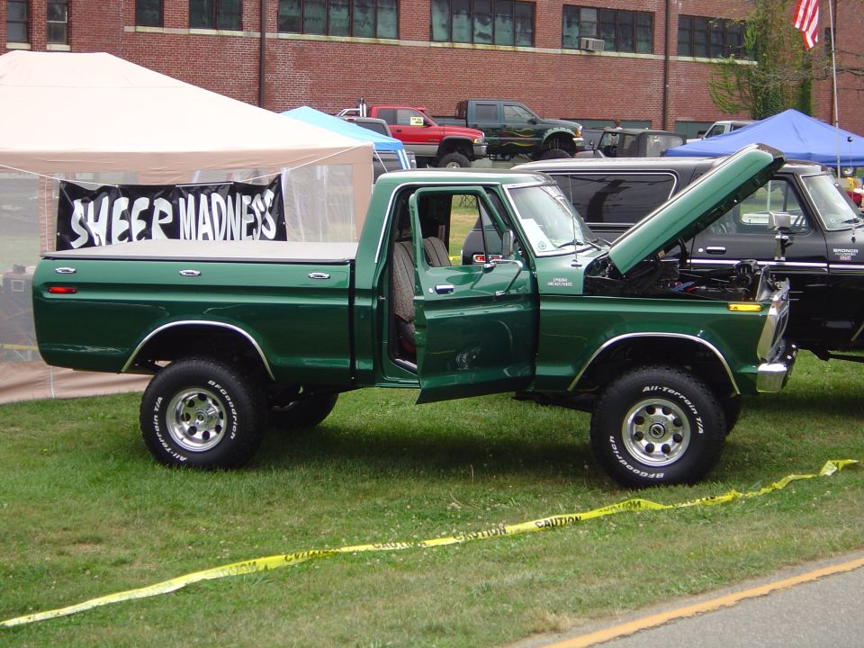 Green pickup truck with hood and door open, parked on grass near tents. Another truck is visible behind it