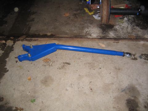 1978 Ford Bronco - A blue-painted tubular radius arm lies on a concrete garage floor, with a forked axle-mount bracket and grease zerks at one end and a heim-…