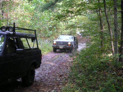 1994 Ford Bronco - Two SUVs navigate a wooded dirt road, with one in the foreground and another approaching.