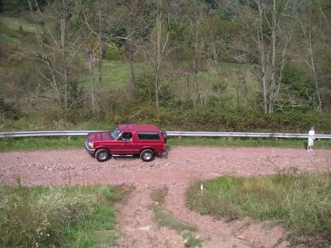 1994 Ford Bronco - A red Ford Bronco drives on a dirt road next to a guardrail, with a person standing nearby.