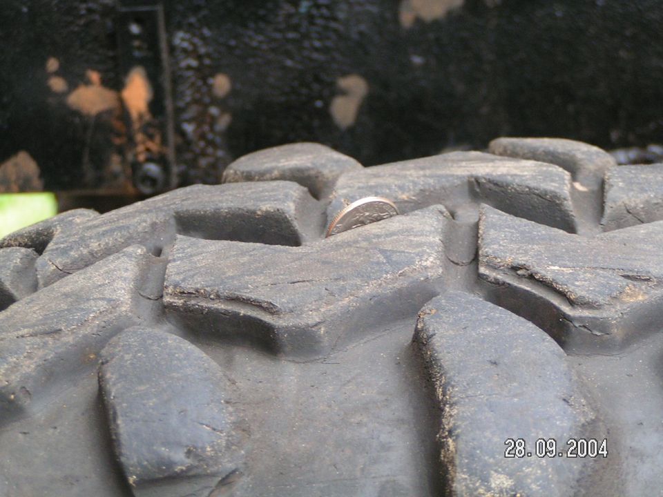 Close-up of a chunky off-road tire tread with a coin in one of the grooves. The date 28.09.2004 is visible