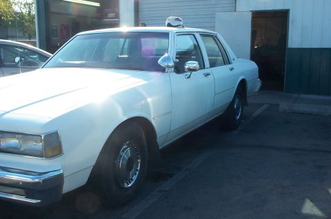 1995 Ford Crown Victoria - A white sedan, likely from the 1980s, is parked in front of a building with an open garage door.