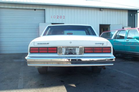 1995 Ford Crown Victoria - Rear view of a white 1980s Chevrolet Caprice sedan parked outside a building, with a taxi nearby.