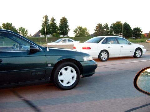1993 Ford Taurus - A dark green sedan is seen on the highway next to a white Ford Taurus.