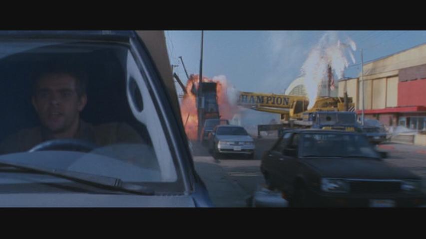 Man driving a car, looking out the window at a large explosion and a crane with the word HAMPION