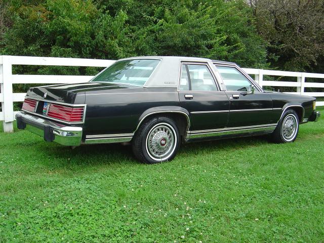 Dark brown Mercury Grand Marquis sedan with tan vinyl top parked on grass by a white fence