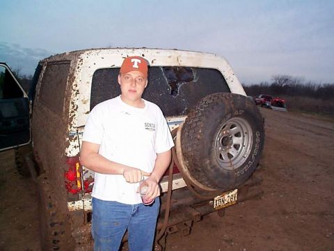 1978 Ford Bronco - A muddy 1978 Ford Bronco is seen from the rear, with a spare tire mounted and a man standing beside it. Other vehicles are visible in the b…