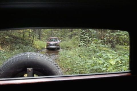 1994 Ford Bronco - View from inside a vehicle looking out at a red SUV driving on a dirt road through a wooded area.