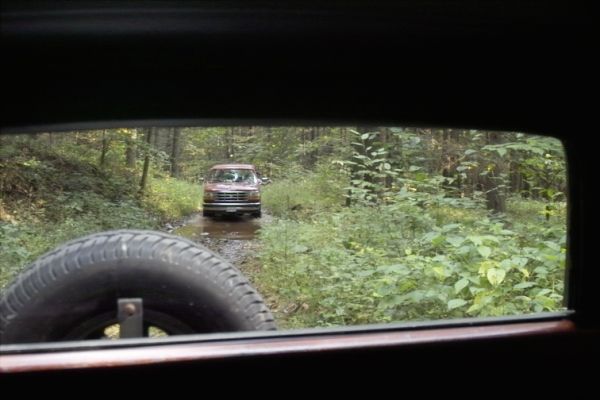 Red SUV on a muddy forest road, seen from behind another vehicle. Spare tire visible in foreground