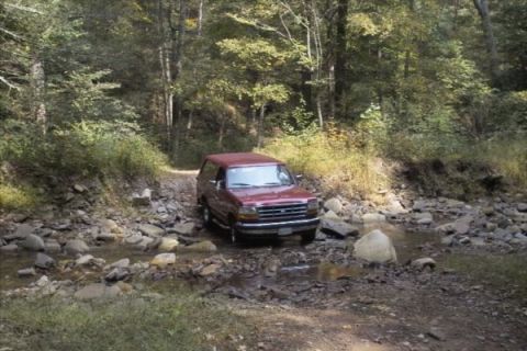 1994 Ford Bronco - A red SUV navigates a shallow, rocky stream bed surrounded by trees.