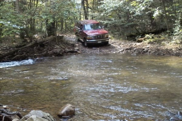 Red 1992 Ford Bronco crossing a stream in a wooded area