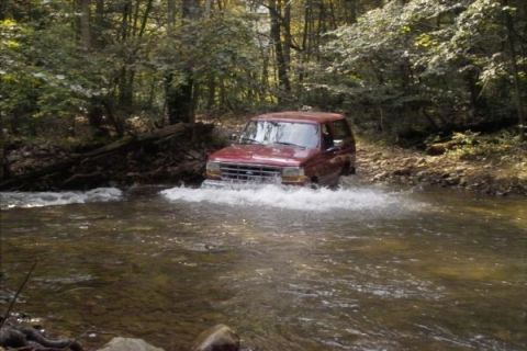 1994 Ford Bronco - A red SUV navigates a shallow creek bed surrounded by dense woods.