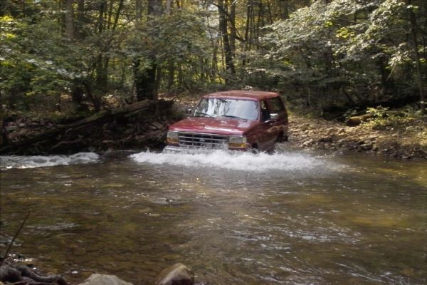 Red SUV driving through a creek, splashing water. Trees and woods in background