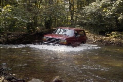 1994 Ford Bronco - A red SUV navigates a stream in a wooded area, kicking up water as it moves forward.