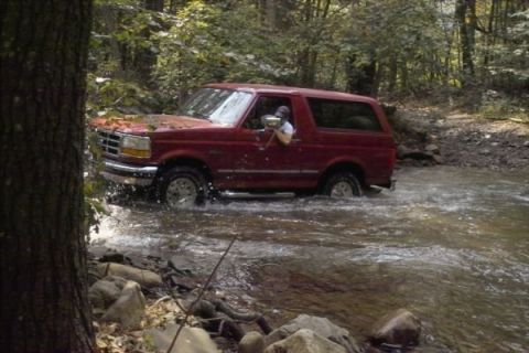 1994 Ford Bronco - A red 1994 Ford Bronco navigates a shallow stream surrounded by trees.
